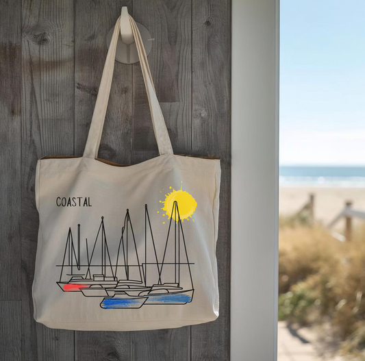 Beige tote bag with sailboat design and 'Coastal' text, hanging on a wooden wall in a cottage with a beach view in the background.