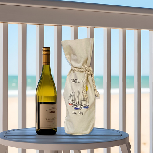 Wine tote with 'coastal wine' text next to a bottle of wine on a table on a deck with the beach in the background