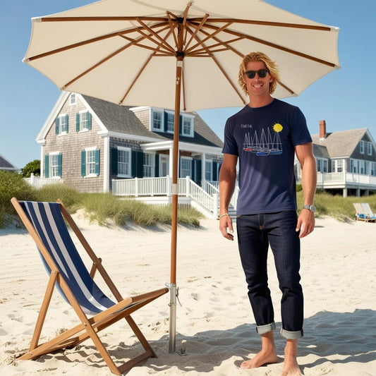 A person standing on a beach in Atlantic Canada under a beige umbrella, wearing a navy t-shirt with coastal line art boat design, sunglasses, and a watch.