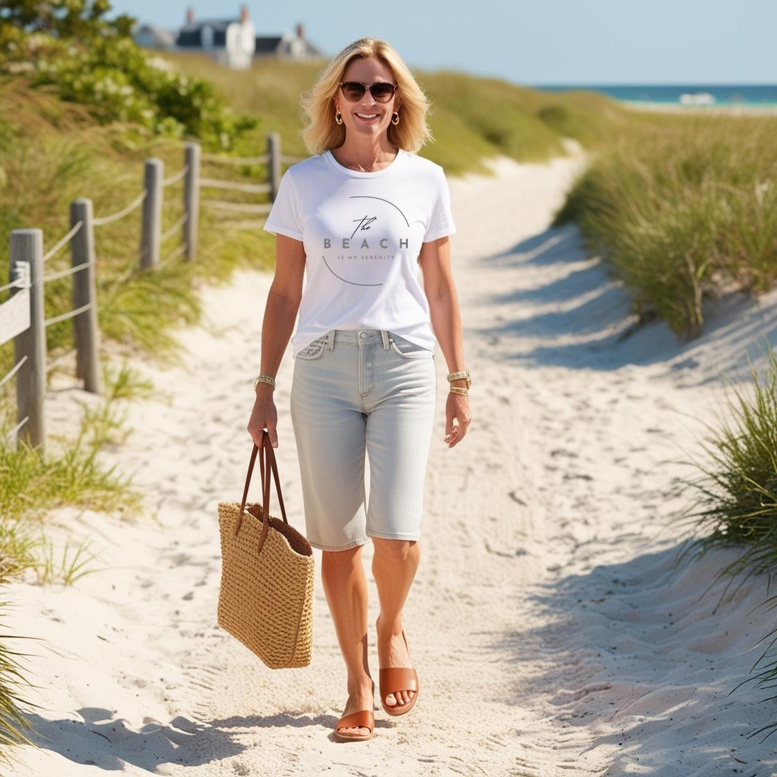 A woman wearing a white t-shirt with the text 'The Beach is my Serenity' on it, paired with light blue denim shorts, sunglasses, and a straw tote bag, walking on a sandy path with beach dunes and grass in the background.
