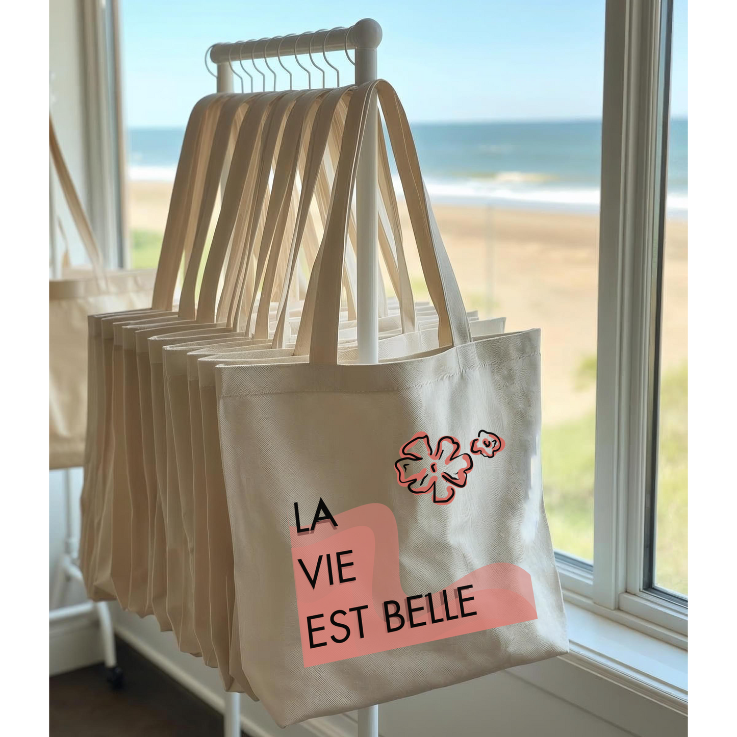 Beige tote bags with 'La Vie Est Belle' design hanging by a window with a beach view.